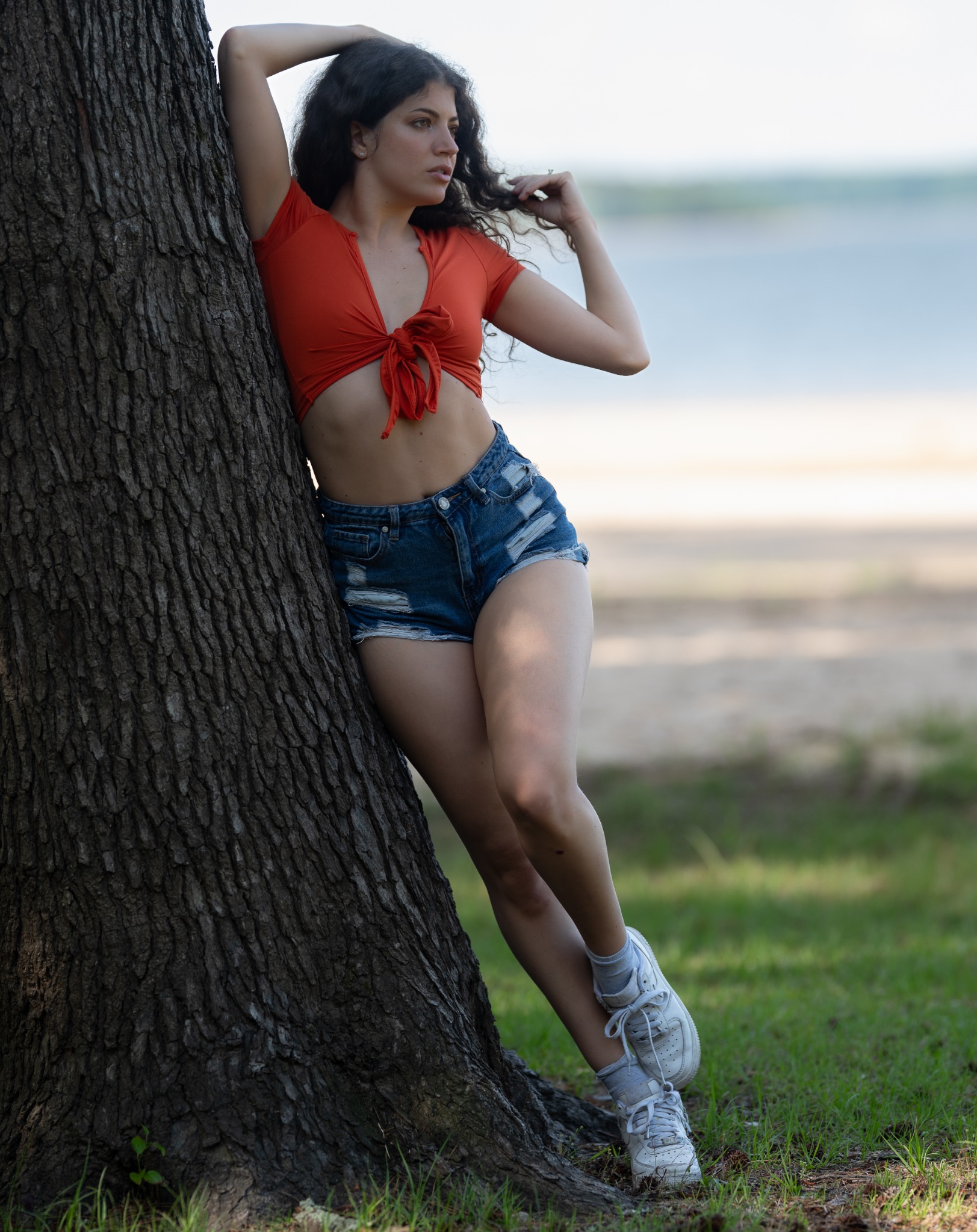Outdoor portrait of a woman in a red top leaning against a lakeside tree